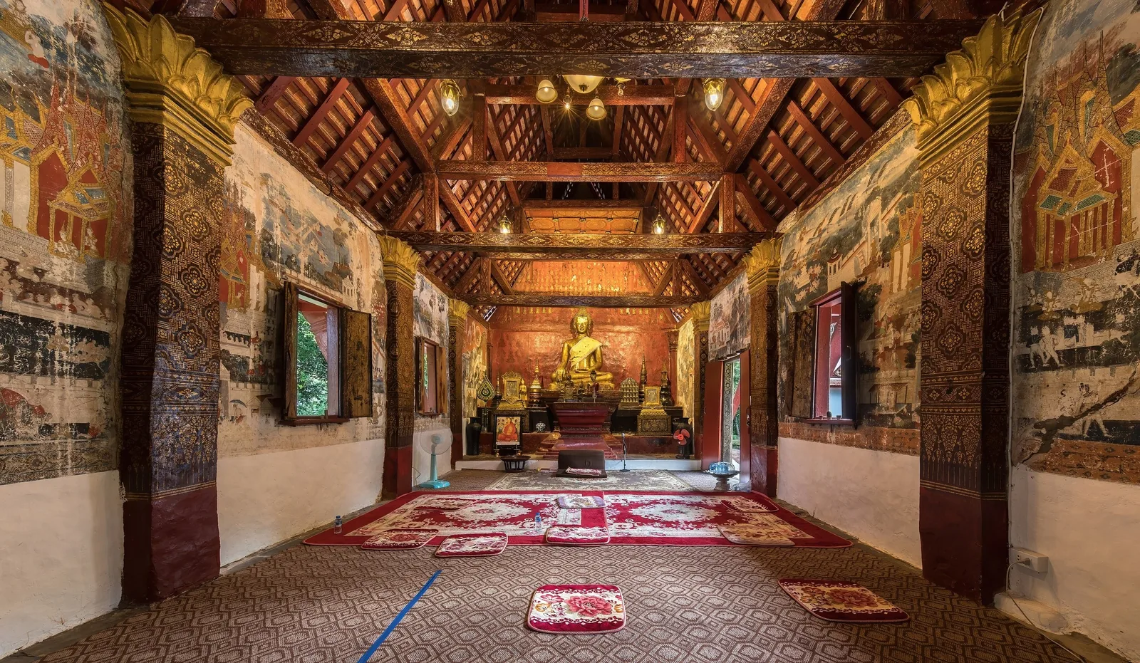 Ornate wooden interior of a temple hall in Luang Prabang with a golden Buddha statue and mural-covered walls.