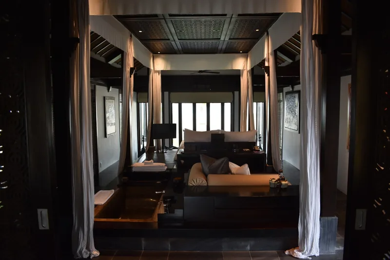 A luxurious dark-wood hotel suite in Hoi An features a canopy bed, sofa, and an in-room bathtub with flowing curtains.