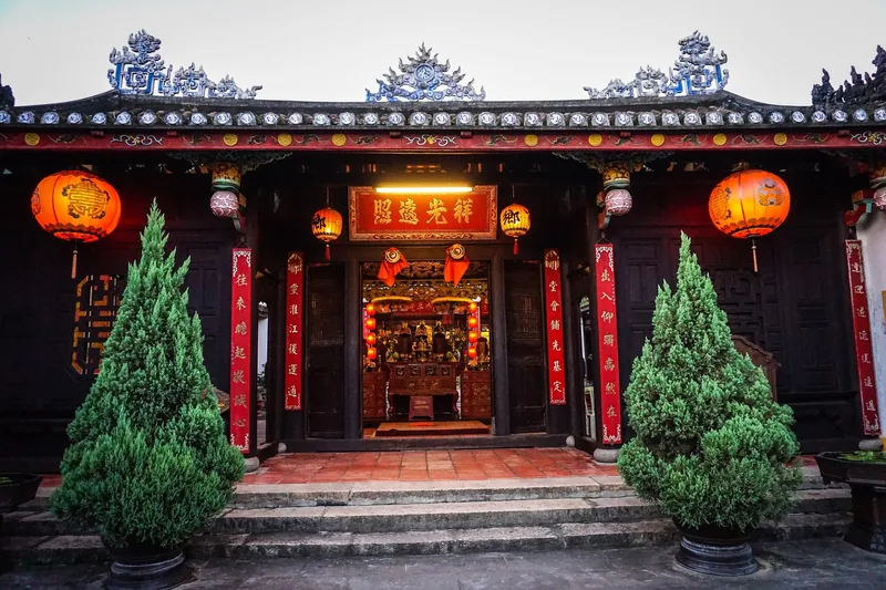 Ornate Chinese assembly hall entrance in Hoi An with red lanterns, carved roof and trimmed potted trees.