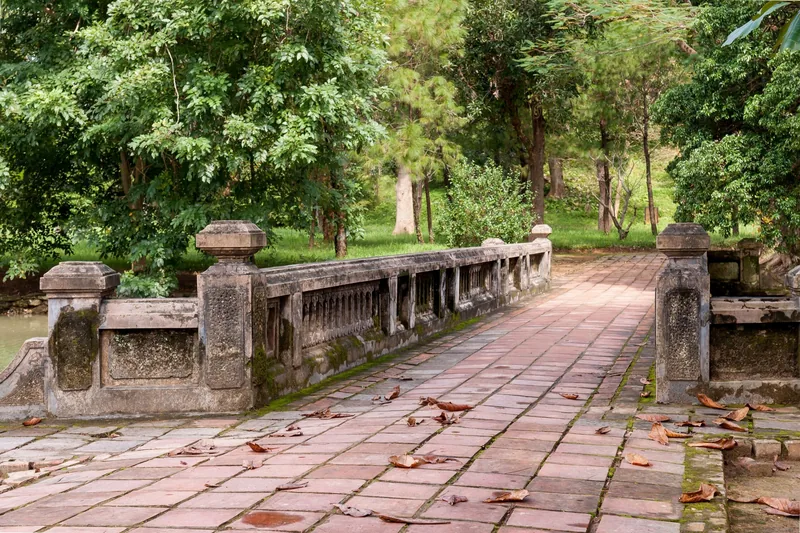 An old stone bridge with weathered balustrades leads through a leafy garden at the Tomb of Emperor Minh Mang in Hue.