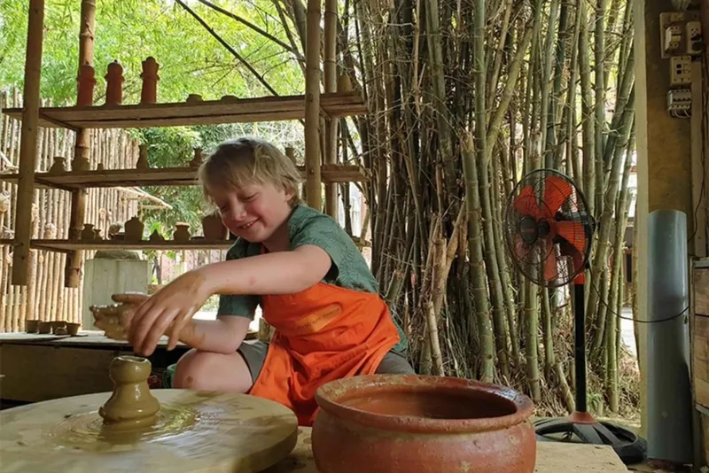 A child in an orange apron shapes clay on a pottery wheel at a traditional craft village in Hoi An.