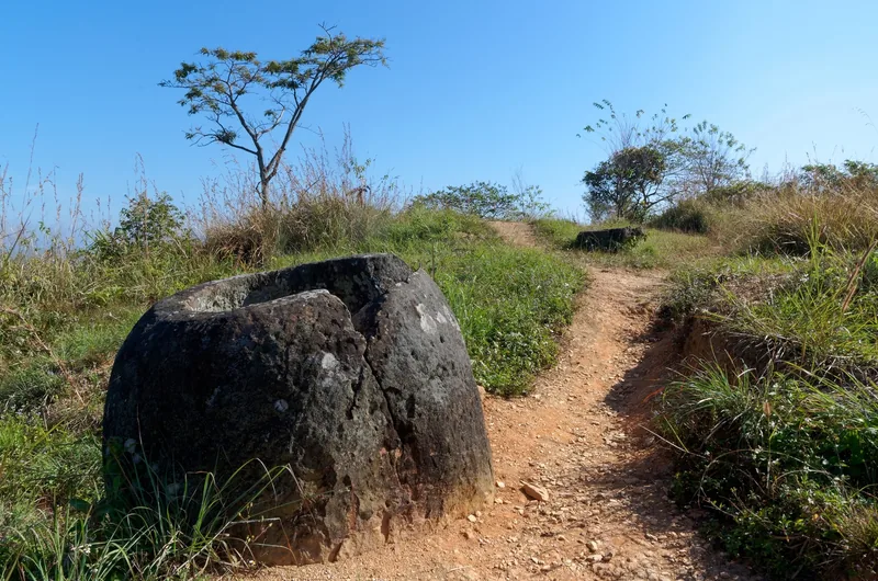 A large stone jar sits beside a dirt path on a grassy hilltop at the Plain of Jars in Laos.