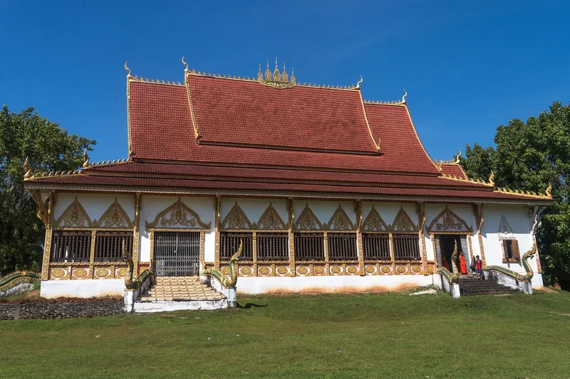 A red-tiled Lao temple with naga staircases stands on a grassy lawn under a clear blue sky in Champasak.