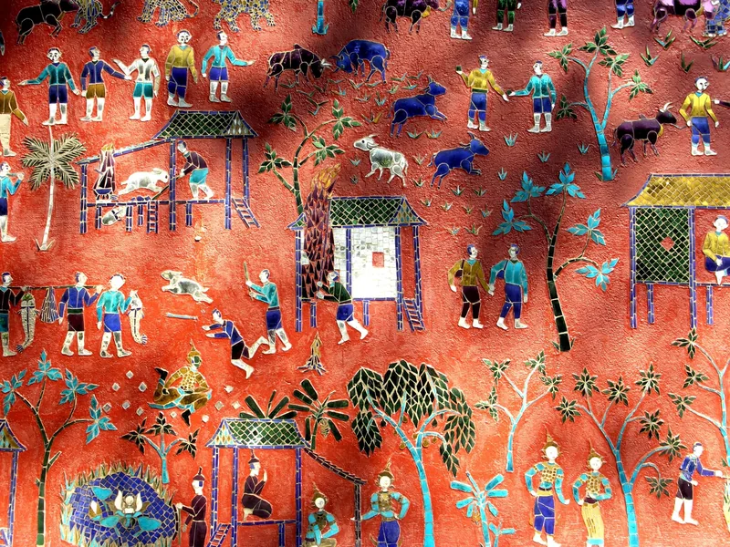 Colorful glass mosaic mural depicting rural village life on a red temple wall at Wat Xieng Thong in Luang Prabang.