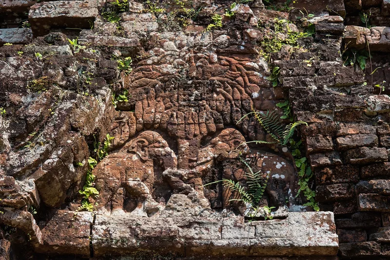 Weathered brick relief carving of an animal figure on an ancient tower wall at My Son, overgrown with ferns