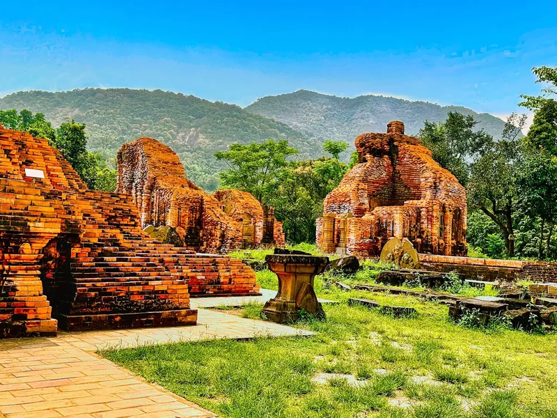 Crumbling red-brick Cham towers of My Son sanctuary stand amid green lawns with forested mountains behind.
